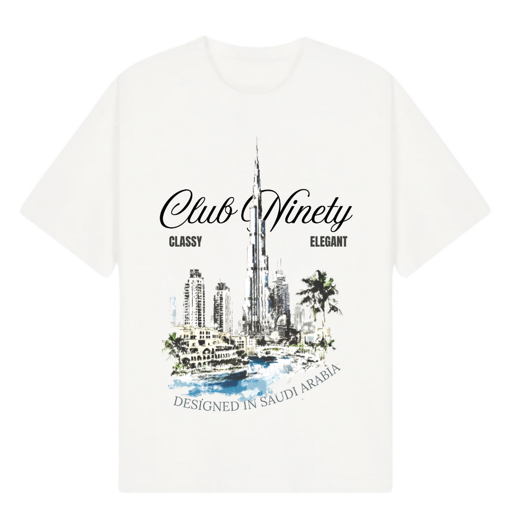 A white short-sleeve t-shirt with a graphic print of the Burj Khalifa in Dubai, featuring the text 'Club Ninety' and 'Classy Elegant'. The image includes promotional text overlays from 'The Gallery Drop' and 'Club Ninety'.