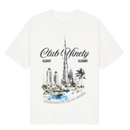 A white short-sleeve t-shirt with a graphic print of the Burj Khalifa in Dubai, featuring the text 'Club Ninety' and 'Classy Elegant'. The image includes promotional text overlays from 'The Gallery Drop' and 'Club Ninety'.