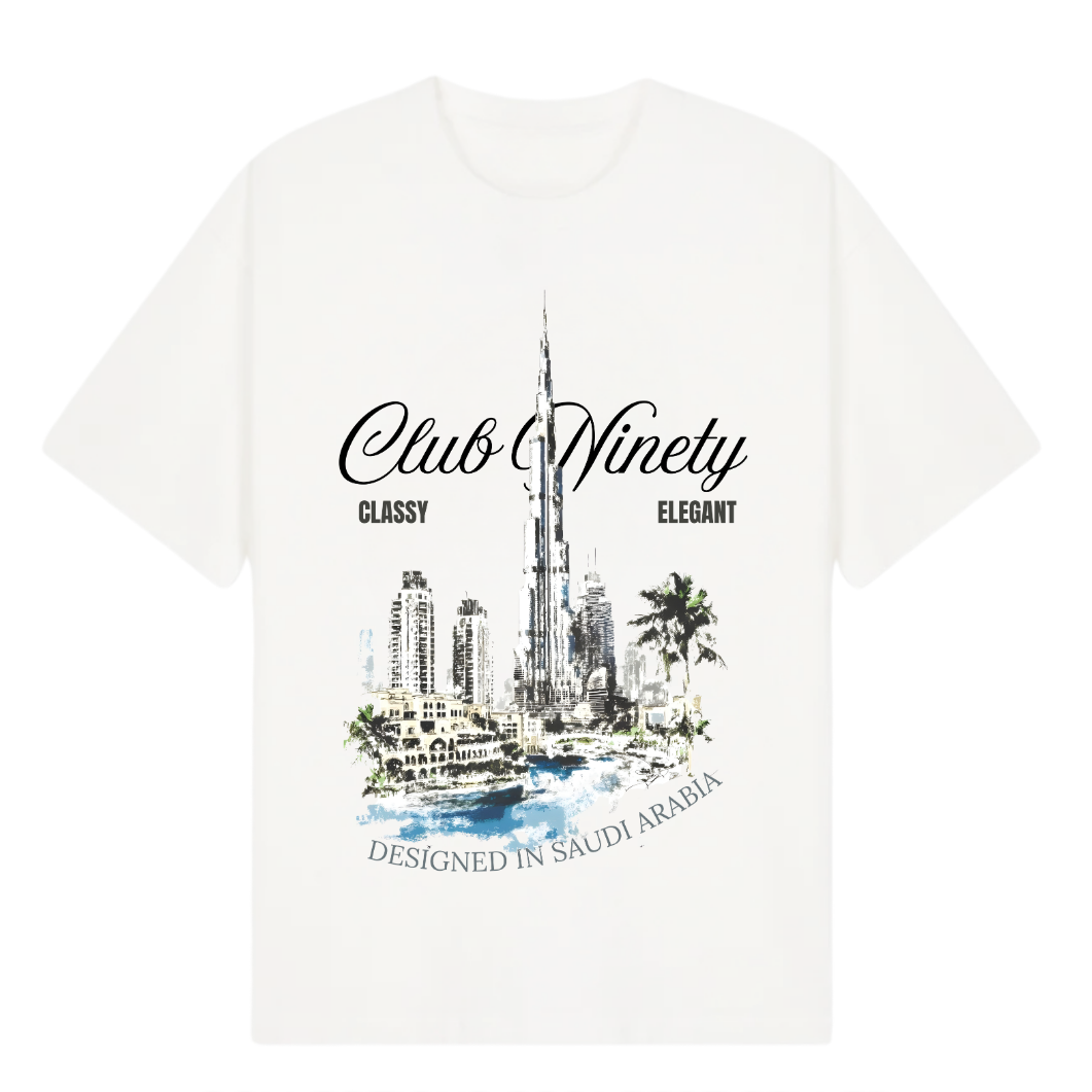 A white short-sleeve t-shirt with a graphic print of the Burj Khalifa in Dubai, featuring the text 'Club Ninety' and 'Classy Elegant'. The image includes promotional text overlays from 'The Gallery Drop' and 'Club Ninety'.