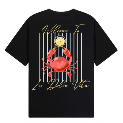 A black short-sleeve t-shirt with a graphic print on the front, featuring a red crab, a clock, vertical blue stripes, and gold script text reading 'Welcome To La Dolce Vita'. The image includes branding text overlays for 'THE GALLERY DROP', 'PEACE COLLECTIVE TEE', and 'CLUB NINETY' on a white background.