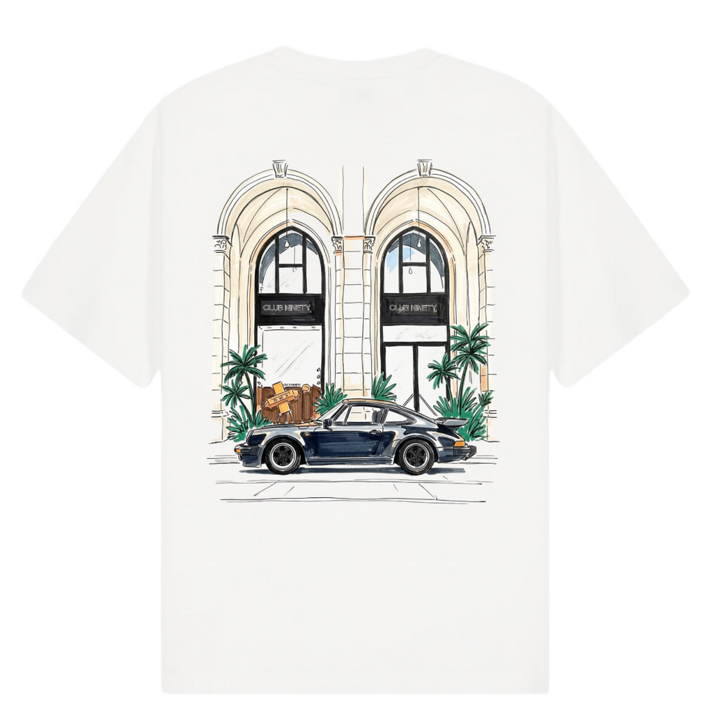 A white short-sleeve t-shirt featuring a colorful graphic print of a vintage blue sports car parked in front of a building with arched windows and palm trees. The image includes text overlays reading 'THE GALLERY DROP LA DOLCE VITA TEE' on the top left, 'CLUB NINETY' on the top right, and 'COLLECTIONS SS26 BEST SELLER' on the bottom.