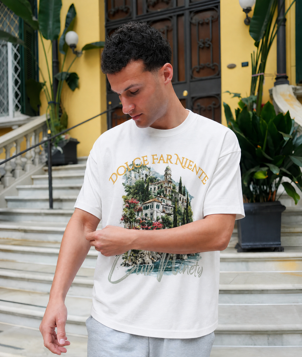 A man wearing a white graphic t-shirt with a vintage-style villa print and the text 'DOLCE FAR NIENTE' and 'Città Venerata', standing on outdoor stone steps in front of a yellow building.