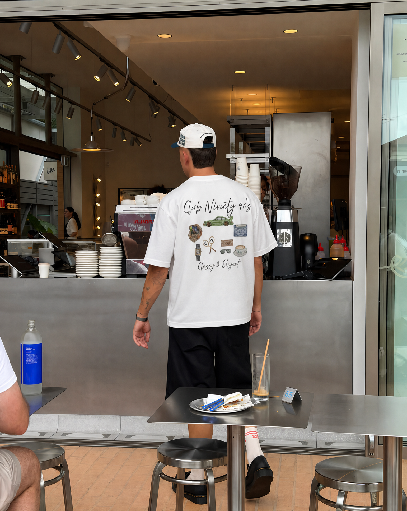 A man standing with his back to the camera in a cafe, wearing a white t-shirt with a graphic print on the back that reads 'Coffee N' Life' and 'Crazy & Bold'.