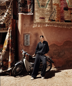 A model wearing a black zip-up hoodie and matching wide-leg sweatpants stands next to a vintage cream-colored scooter in front of a rustic terracotta wall with hanging rugs.