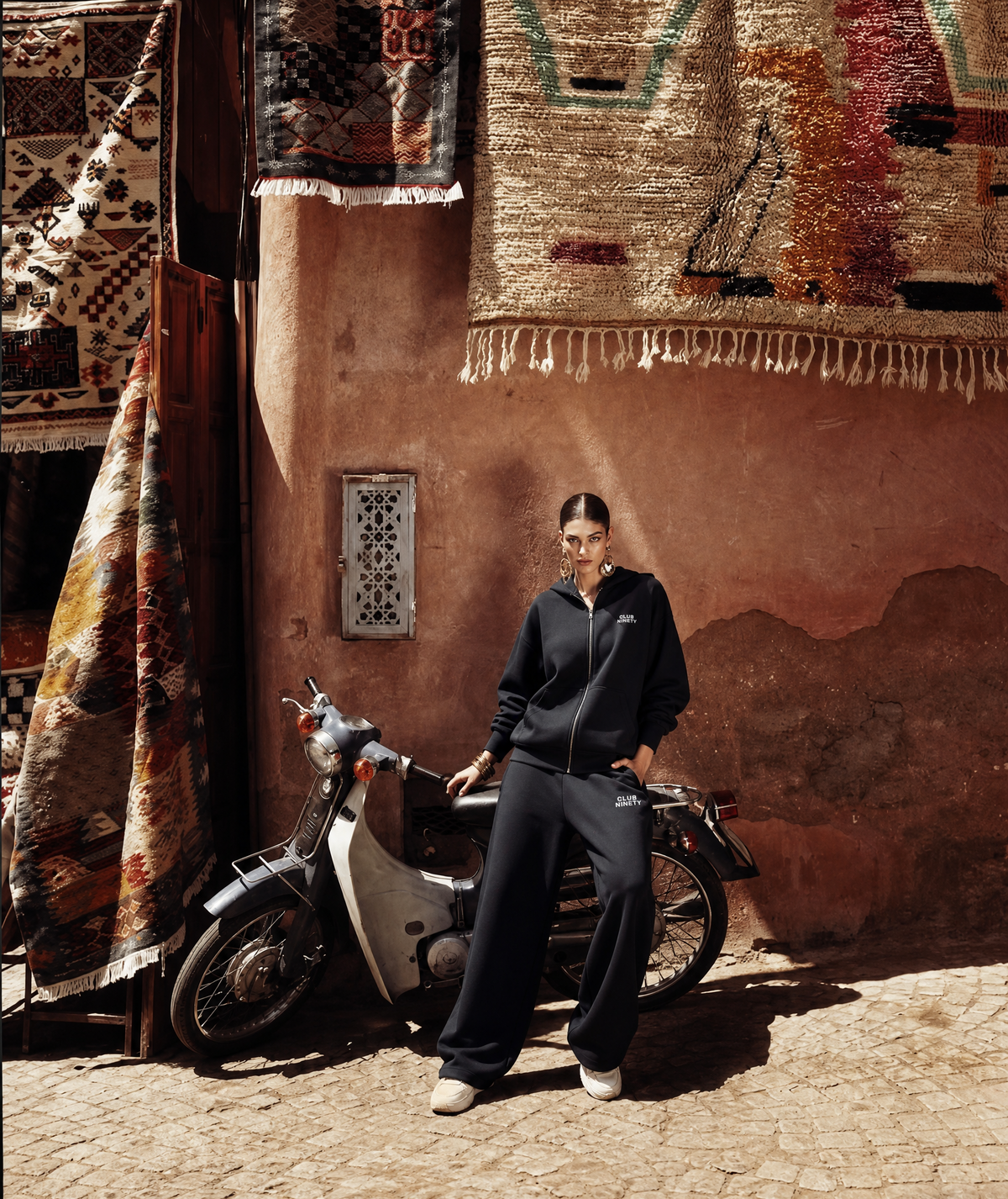 A model wearing a black zip-up hoodie and matching wide-leg sweatpants stands next to a vintage cream-colored scooter in front of a rustic terracotta wall with hanging rugs.