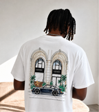A man viewed from behind wearing a white graphic t-shirt with a detailed illustration of a vintage car parked in front of a building with arches, paired with light blue jeans in a minimalist indoor setting.