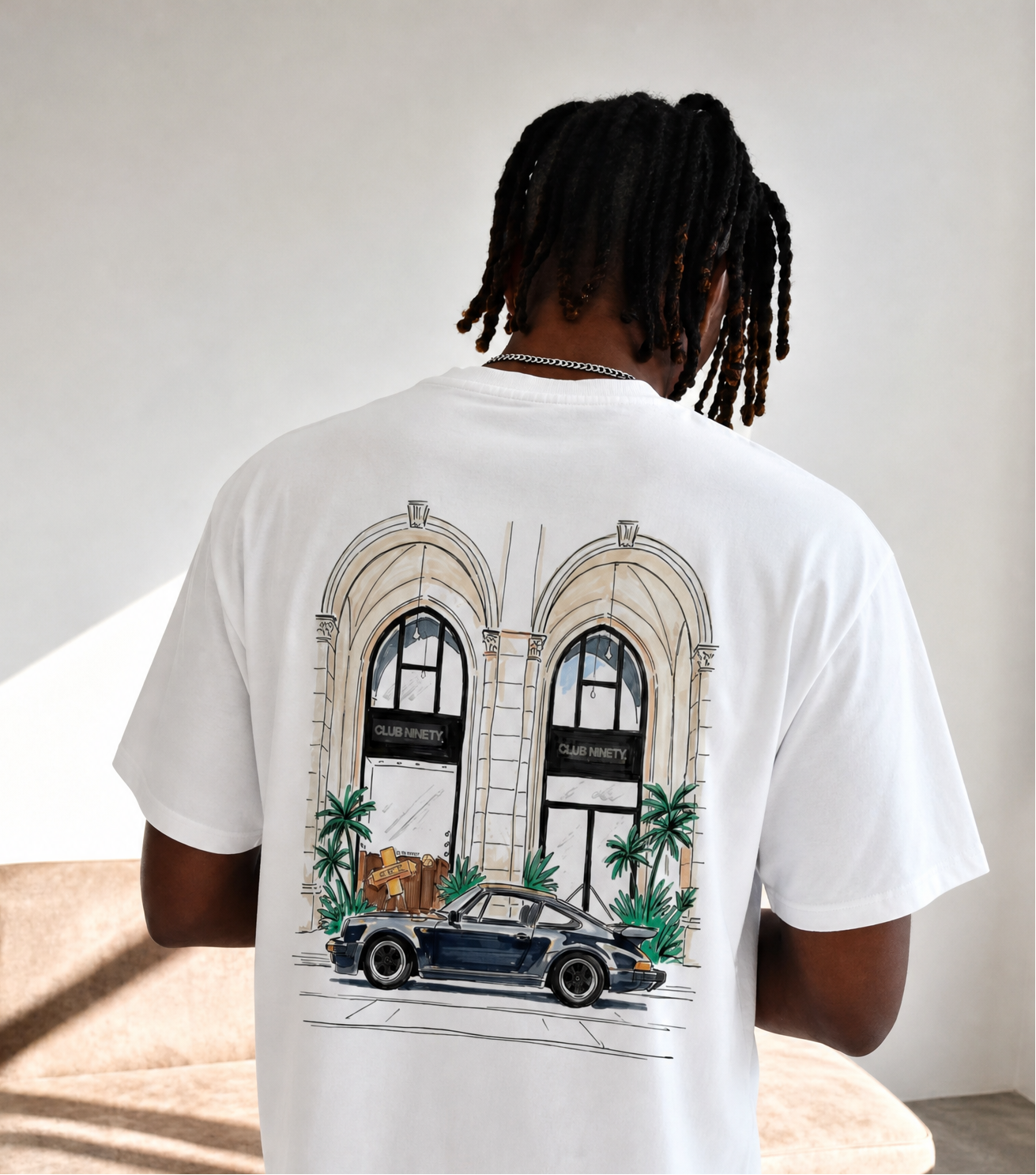 A man viewed from behind wearing a white graphic t-shirt with a detailed illustration of a vintage car parked in front of a building with arches, paired with light blue jeans in a minimalist indoor setting.