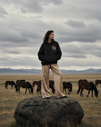 A model stands on a large rock in an open grassland under a cloudy sky, wearing a black hoodie with a horse graphic and beige wide-leg pants. Several horses graze in the background, with mountains visible in the distance.